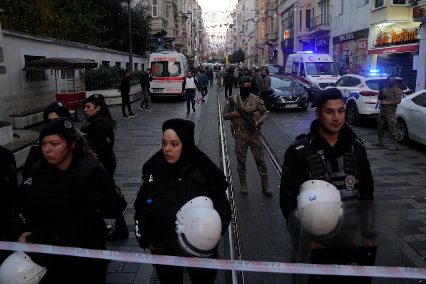 Security and military personnel guard a street while ambulance crews work behind them.