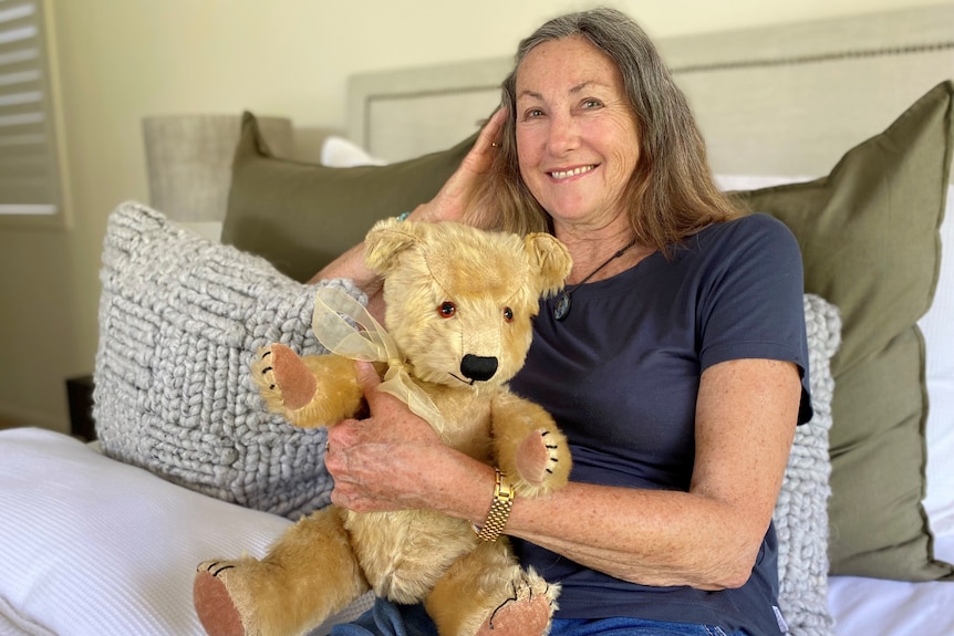 Woman sits on a bed with Steiff bear, smiling