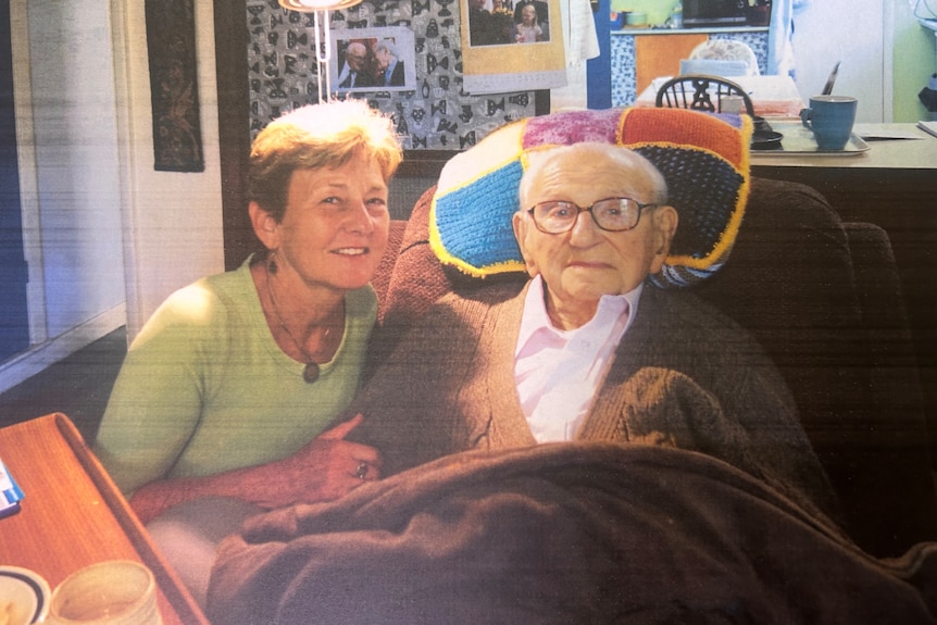 A woman smiles next to a frail man wearing glasses on a couch.