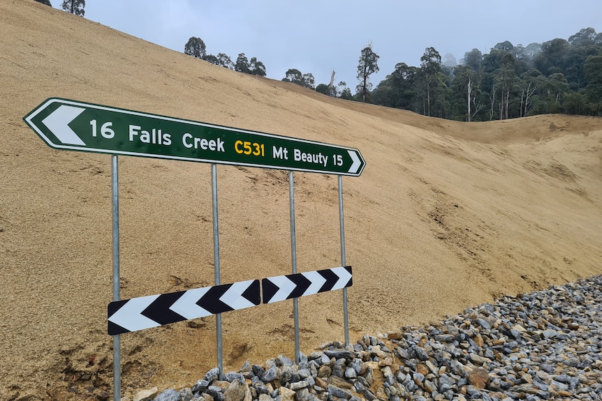 A sandy slope with a wide rock base lies at the road sign pointing to Falls Creek and Mount Beauty.