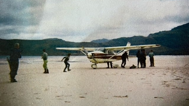 A white light aircraft waiting on the white sands of Lake Pedder, surrounded by several people and their walking equipment. 