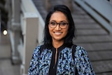 A smiling woman with dark skin, black hair and black-framed glasses, wearing a blue patterned top