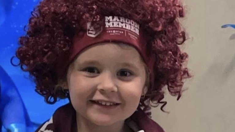 A toddler wearing Queensland Maroons rugby league merchandise smiles at the camera. 