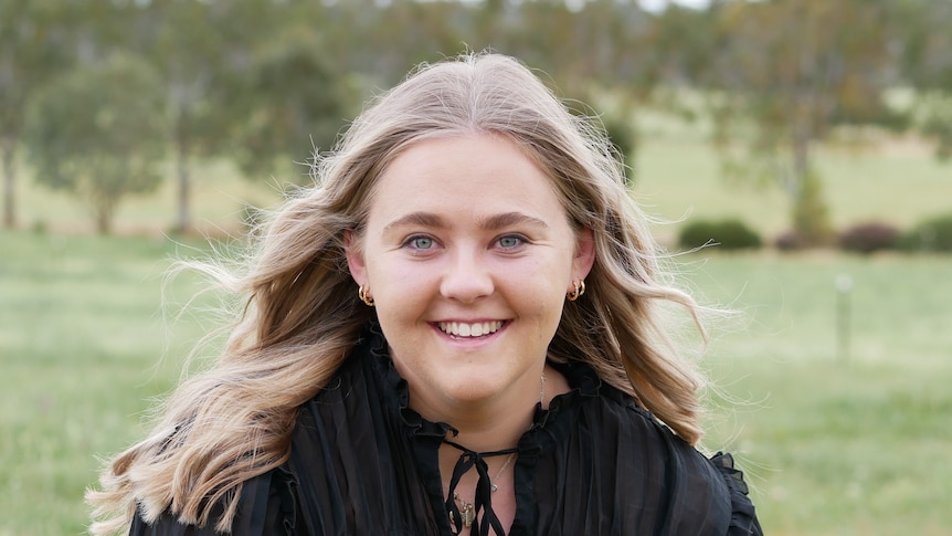 Portrait of a blonde girl wearing a black shirt, with a paddock in the background. 
