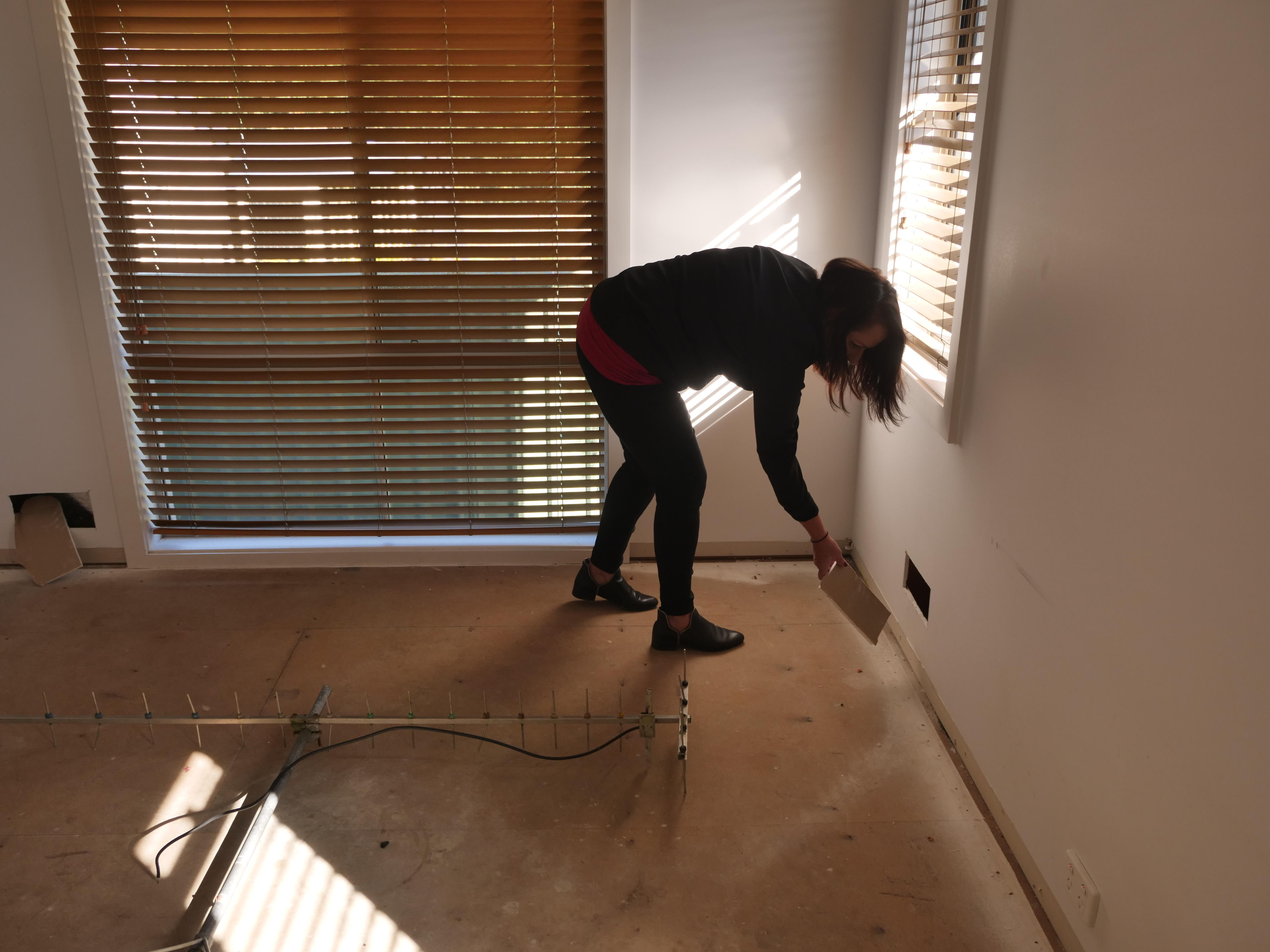 A woman leans over to pick up a piece of plaster cut from a wall in an empty house with wooden floorboards.