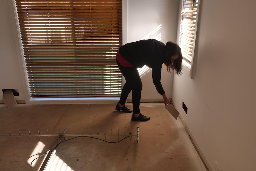 A woman leans over to pick up a piece of plaster cut from a wall in an empty house with wooden floorboards.