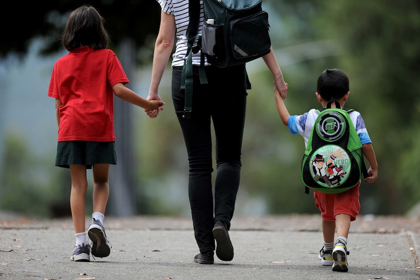 A mother walks holding the hands of two children on their way to school holding backpacks.