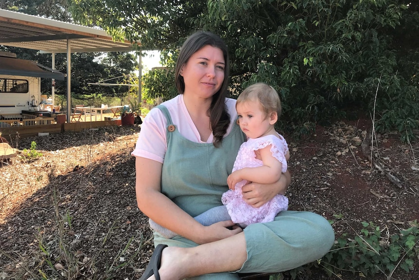 Woman with long brown hair sitting in front of a tree outside her home, with her daughter Warner on her lap