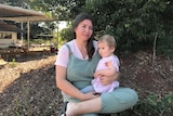 Woman with long brown hair sitting in front of a tree outside her home, with her daughter Warner on her lap