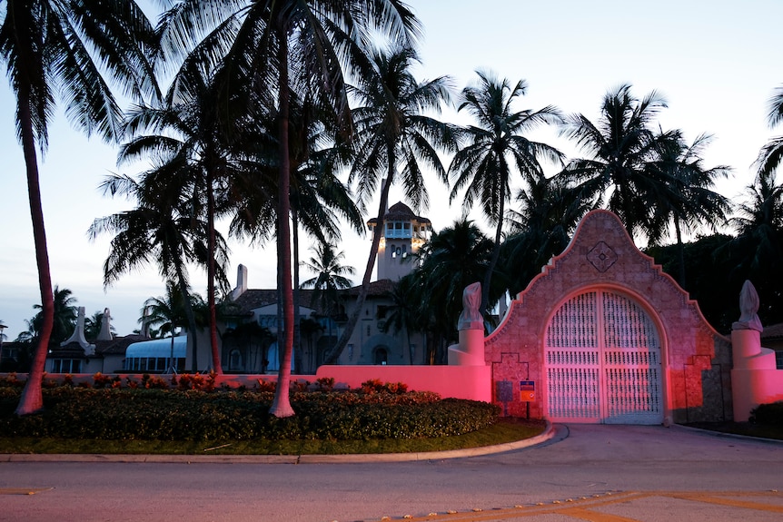A grand facade entrance to an estate lined with palm trees is lit up in red lights from a law enforcement car.
