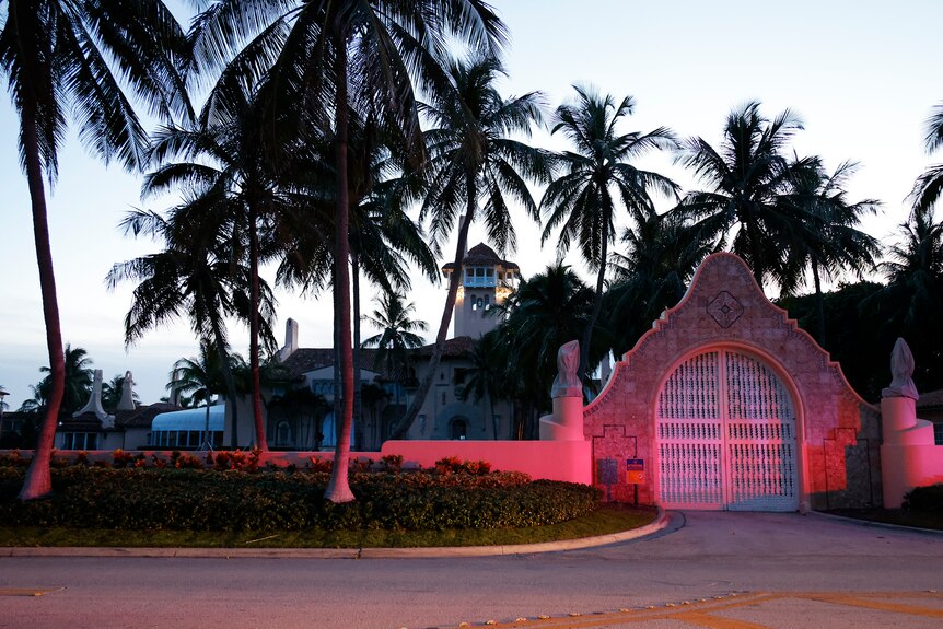 A grand facade entrance to an estate lined with palm trees is lit up in red lights from a law enforcement car.