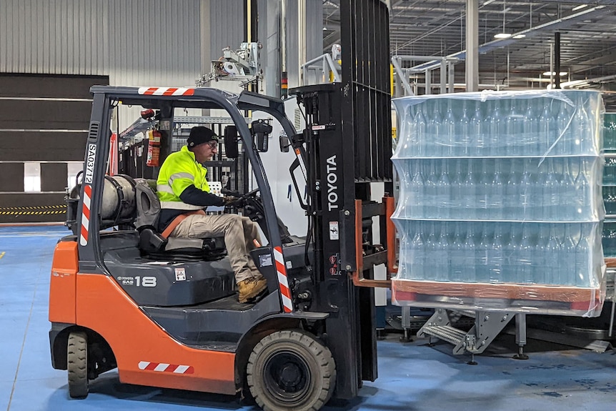 A man in a high vis top driving a forklift, lifting a pallet of glass wine bottles.