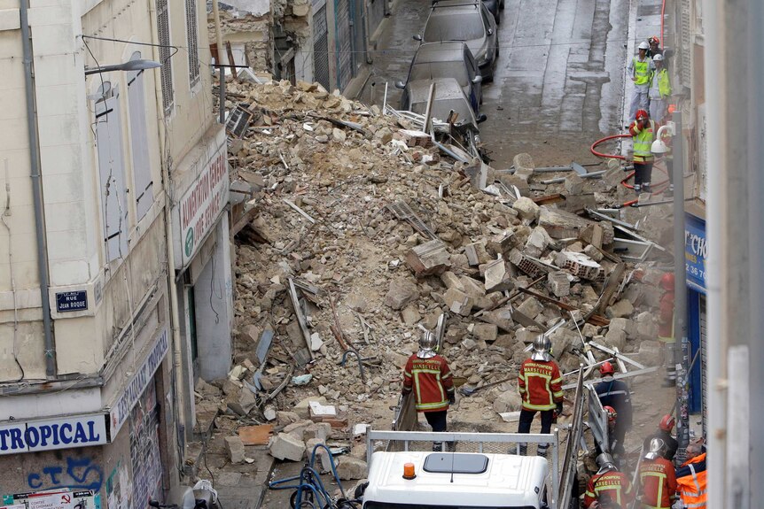 A large pile of rubble spilled out on to a narrow street, with dust covering cars parked nearby.