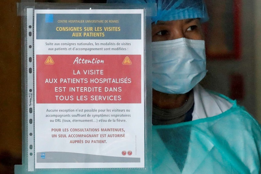 A health worker wearing a hospital mask looks out from a window behind a coronavirus warning sign