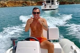 A shirtless man gives the thumbs up while driving a boat.