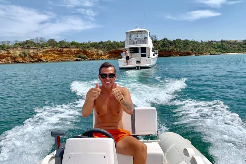 A shirtless man gives the thumbs up while driving a boat.