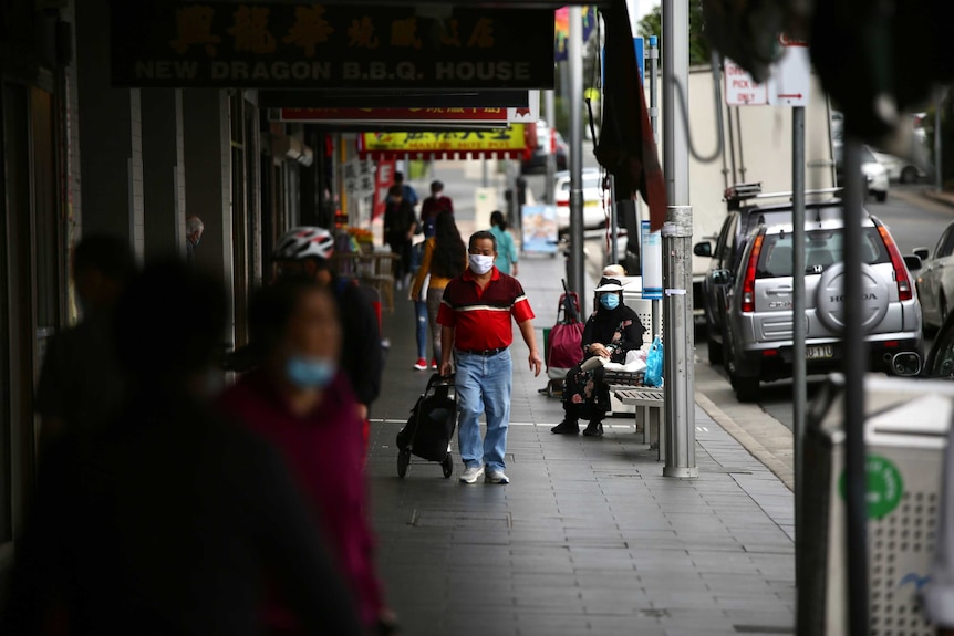 A number of people wear face masks as they walk on the path of a strip mall in Auburn. A woman sits.