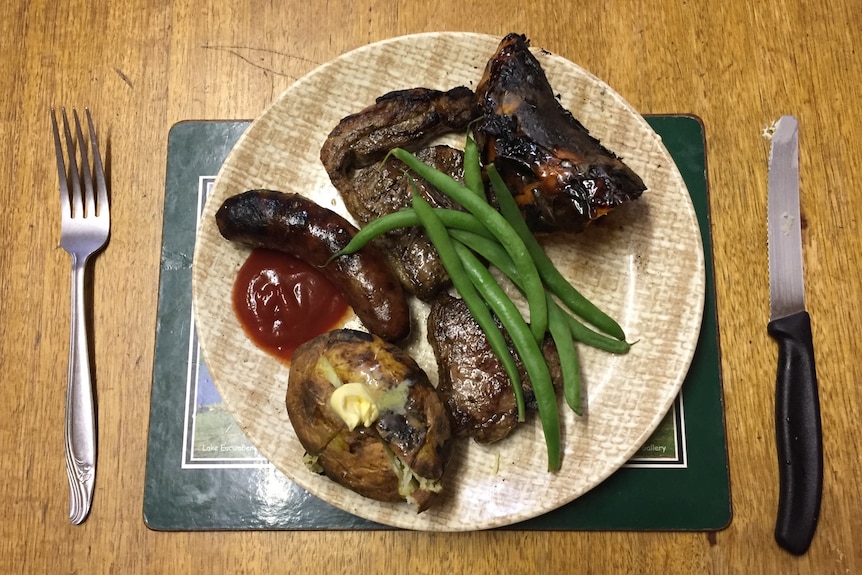 Une assiette et des couverts avec des côtelettes d'agneau grillées, des saucisses, des pommes de terre et des haricots verts.