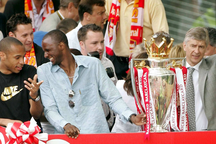 Casually dressed soccer players celebrate as their manager holds the Premier League trophy.