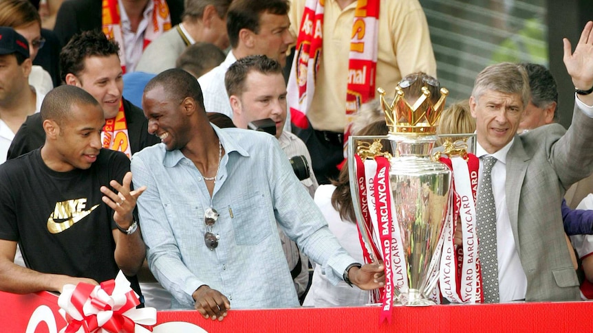 Casually dressed soccer players celebrate as their manager holds the Premier League trophy.