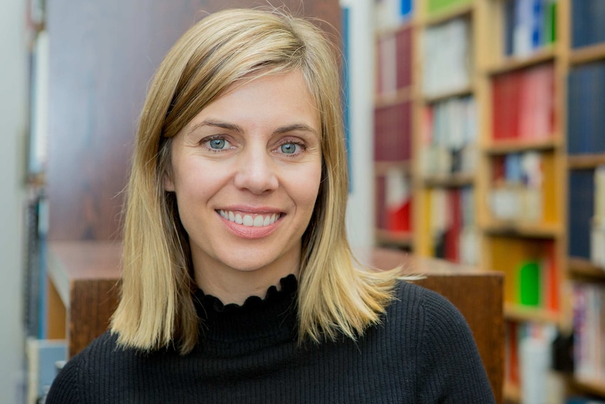 A blonde woman smiles at the camera with a bookshelf behind her