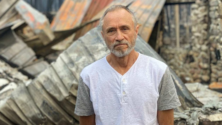 A man stands in front of the shell of a stone-built house that was destroyed by bushfire.