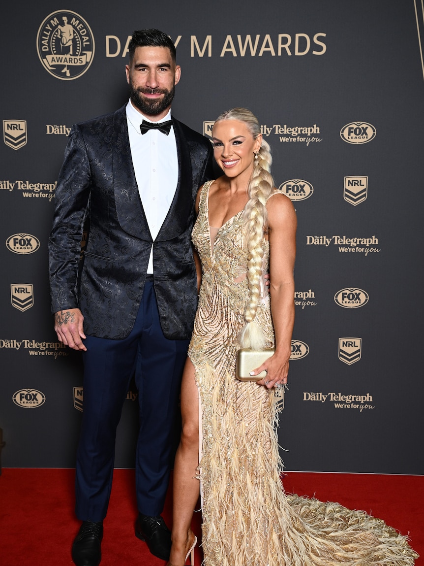 James wears a suit and poses next to his partner who is wearing a long gold beaded dress. 