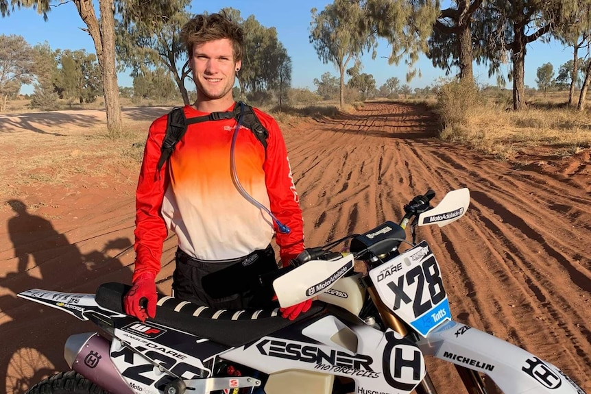 A man wearing an orange shirt stands with his white dirt bike on a red dirt road with gum trees in the background