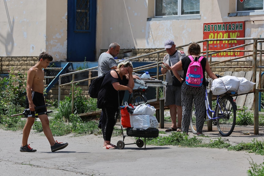 A woman holds her head with a small trolley of belongings outside a building