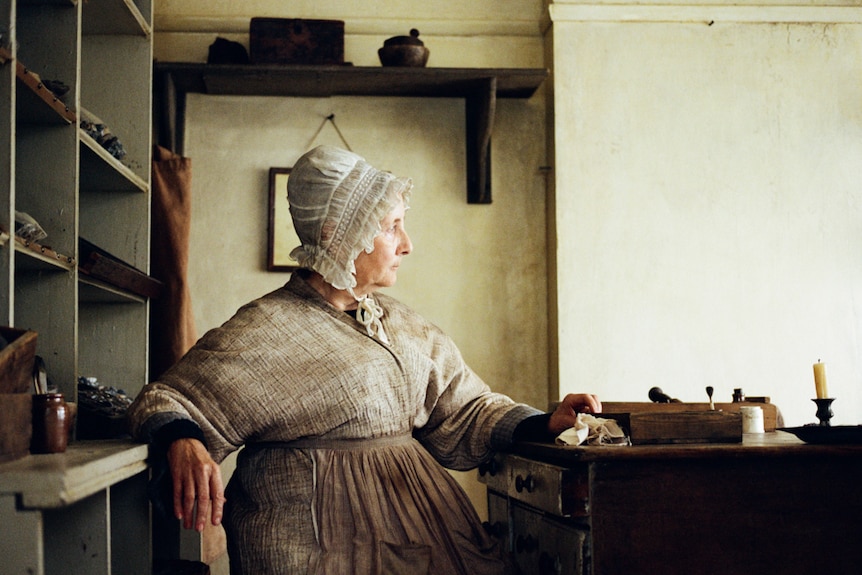 Side profile shot older actress in 19th-century working-class house dress, sitting at kitchen table, staring as if out window.