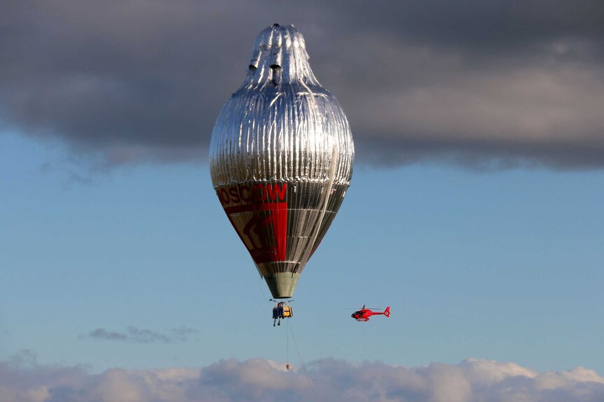 A large silver hot air balloon in the sky with a small red helicopter beside it.
