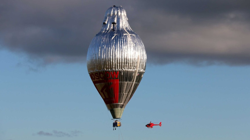 A large silver hot air balloon in the sky with a small red helicopter beside it.