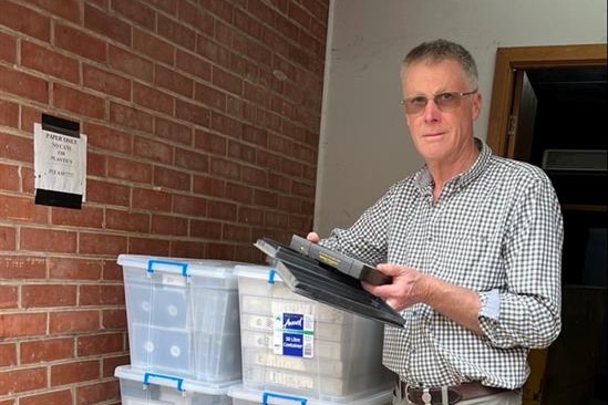 Man in checkered long sleeve shirt and glasses stands next to a pile of plastic boxes filled with historical footage