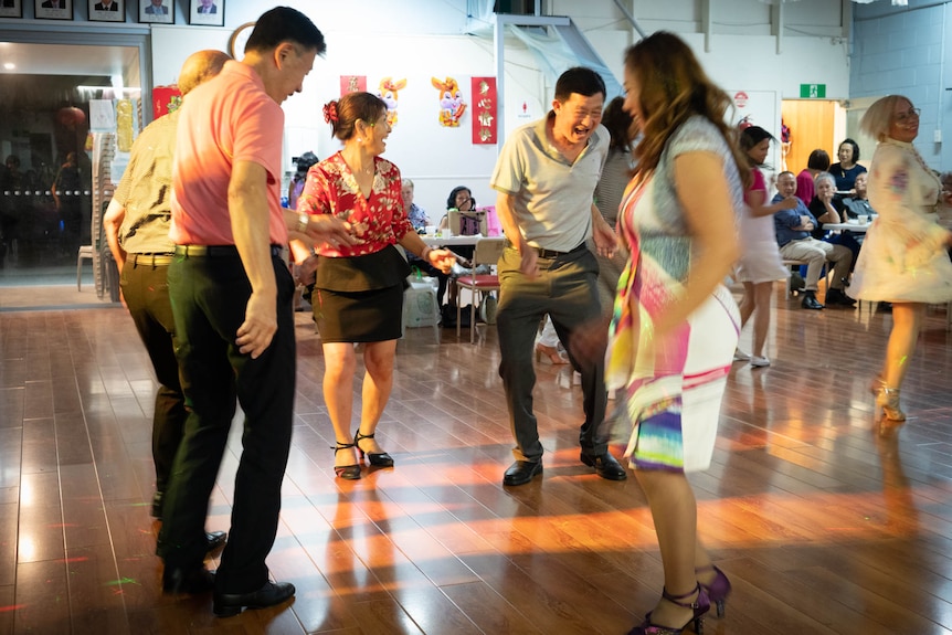 Inside a hall, a group of people laughing and smiling dances. Others are seated behind them.