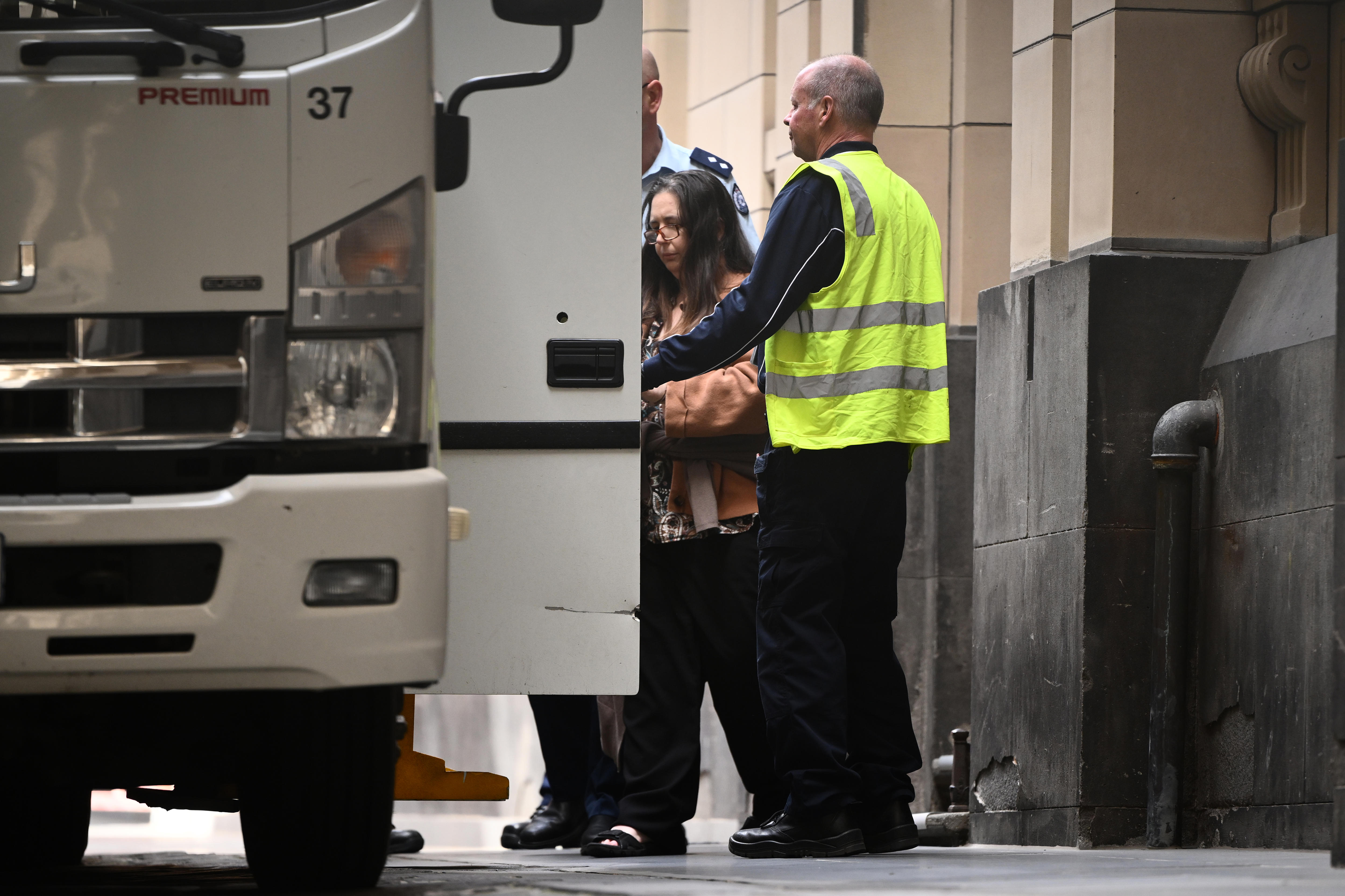 A woman with long dark hair in a brown top is escorted by security from a sandstone building to a white van.