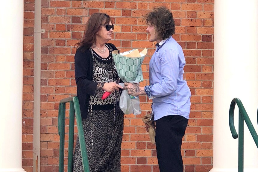 A man hands over a bunch of flowers to a woman with long brown hair and sunglasses.