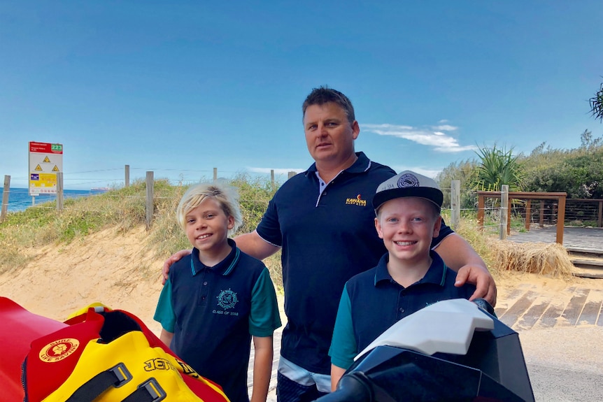 A man stands with his arms around two 11-year-old boys at the beach.