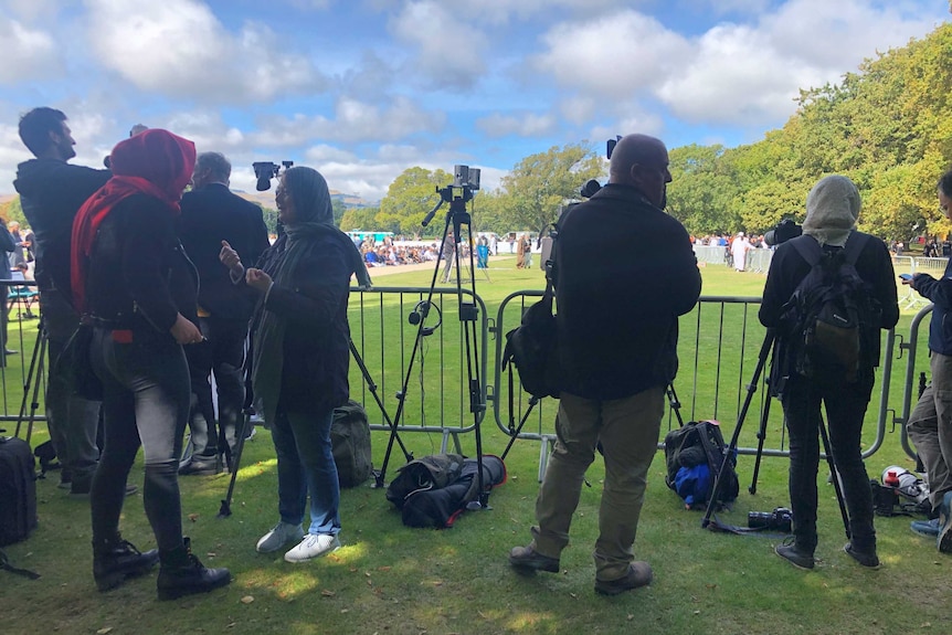 Wide shot of media and cameras on tripods set up at park with female journalists and photographer wearing head scarves.