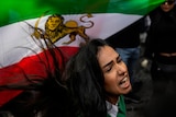A woman screams out, her hair flowing behind her, in front of an Iranian flag with the lion and sun symbol