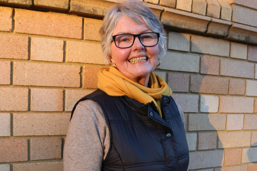 Leonora smiles, standing next to a brick building, wearing a scarf and jacket.