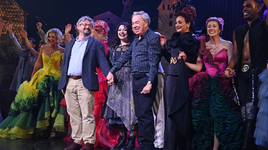 Laurence Connor (left) and Andrew Lloyd Webber (right) on stage holding hands and smiling, flanked by performers in costume.