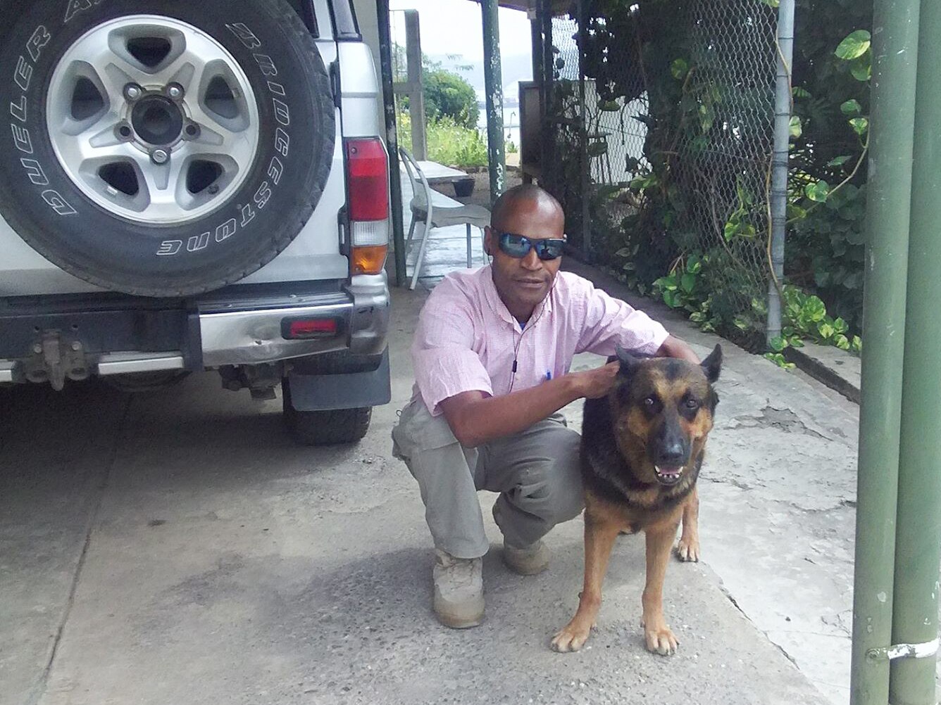 A man in sunglasses and a pink shirt bends down to wrap his arm around a handsome German Shepherd