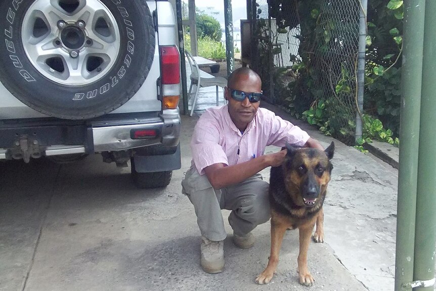 A man in sunglasses and a pink shirt bends down to wrap his arm around a handsome German Shepherd