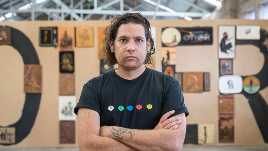 The artist, with his curly brown hair swept back and wearing a black graphic t-shirt, stands in front of his art work OTHER.