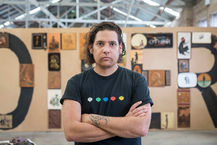 The artist, with his curly brown hair swept back and wearing a black graphic t-shirt, stands in front of his art work OTHER.