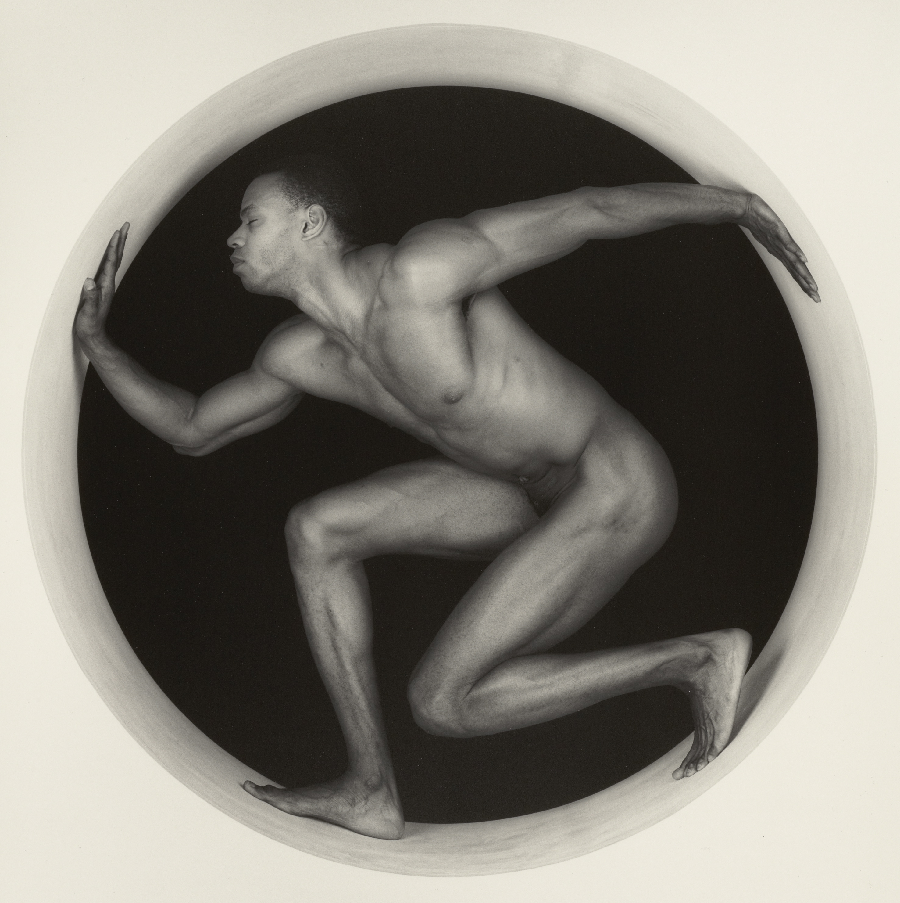 Black and white photograph a muscular nude man with eyes closed standing within a structure with a circular cut out.