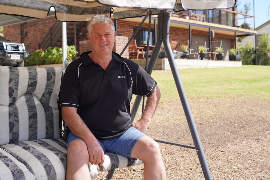 A man wearing a polo shirt and shorts sits on a swinging loveseat amid lawn