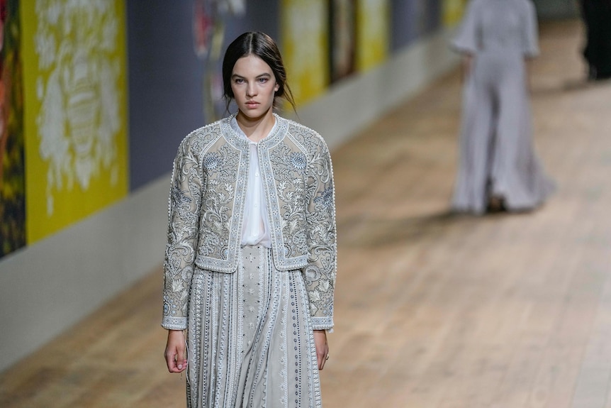 A model wearing a grey and blue embroidered jacket and pants with white shirt. 