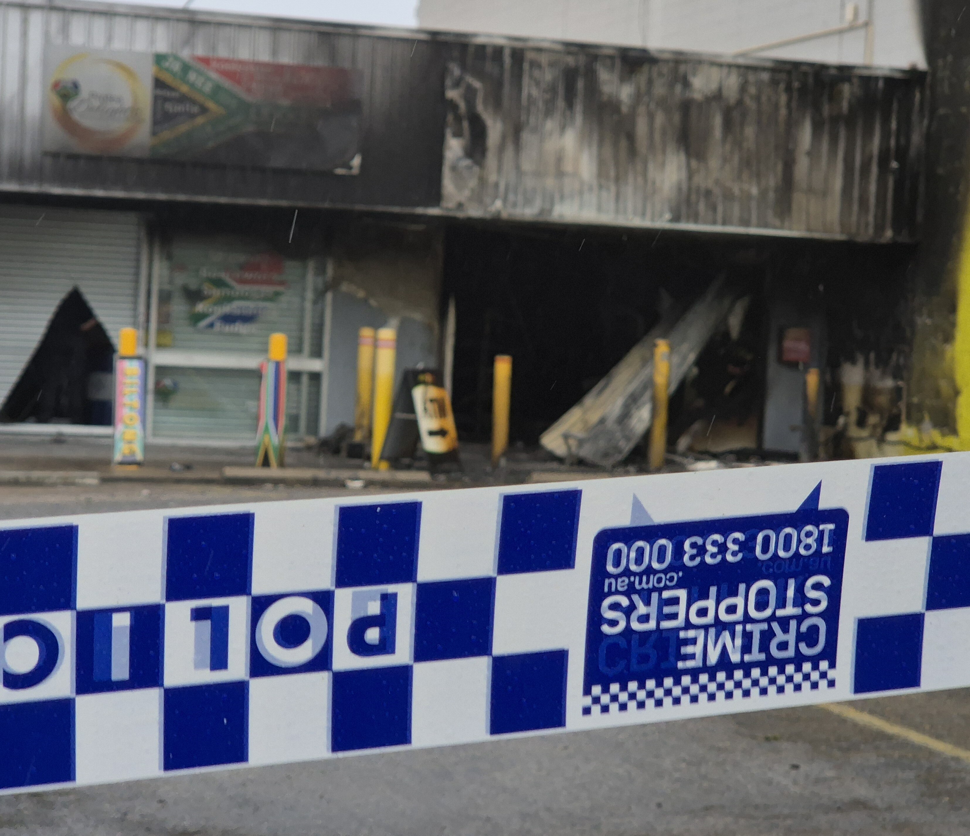Blue and white police tape in front of a burnt shop front which has an ATM sign outside.