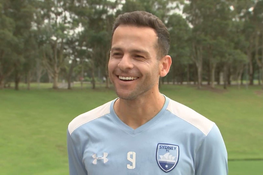 A footballer wearing a Sydney FC shirt stands smiling in a park.
